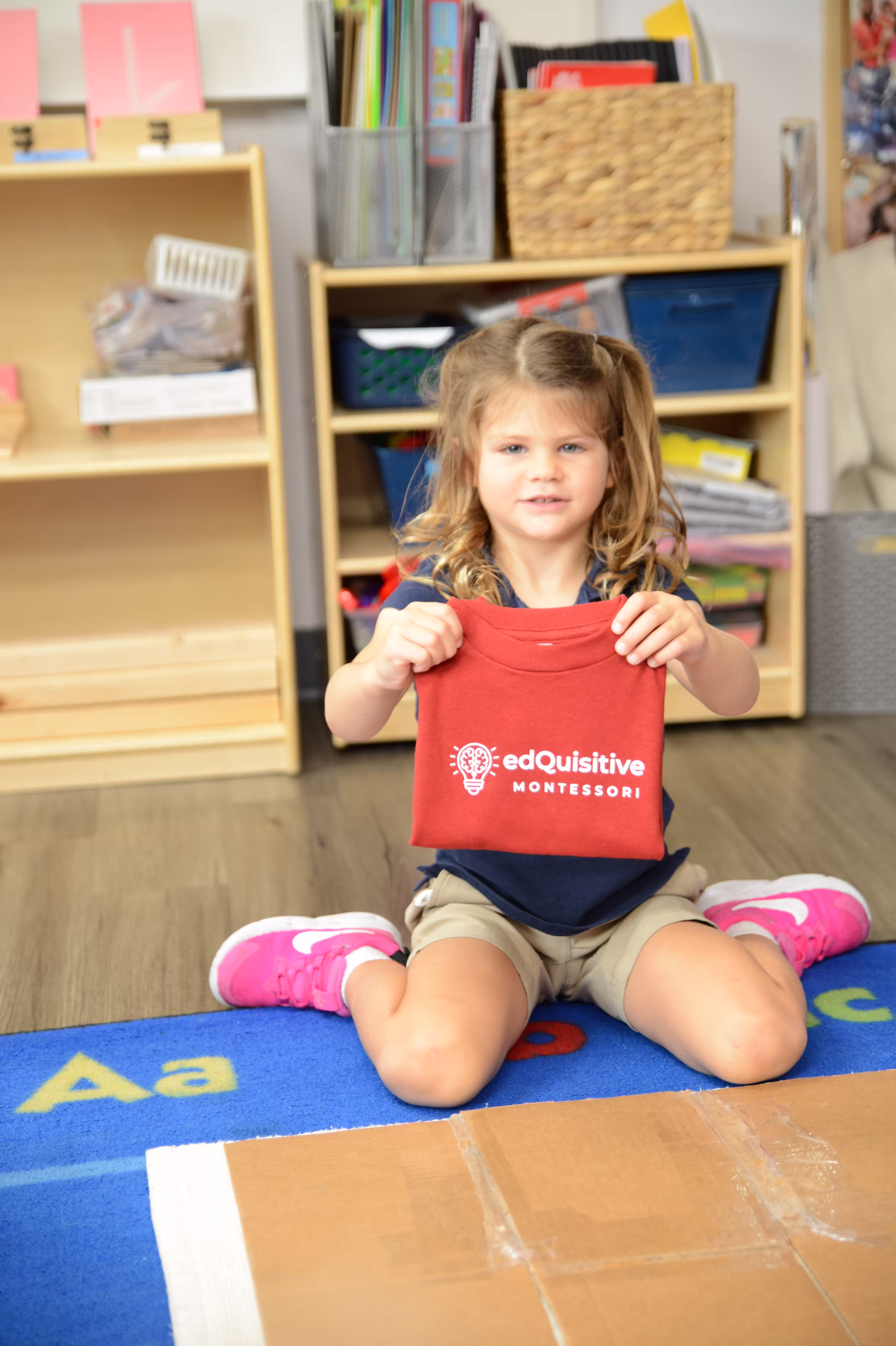 Practical life: folding her own shirt A preschool girl kneeling on a rug holds up a red Edquisitive Montessori shirt she folded herself.