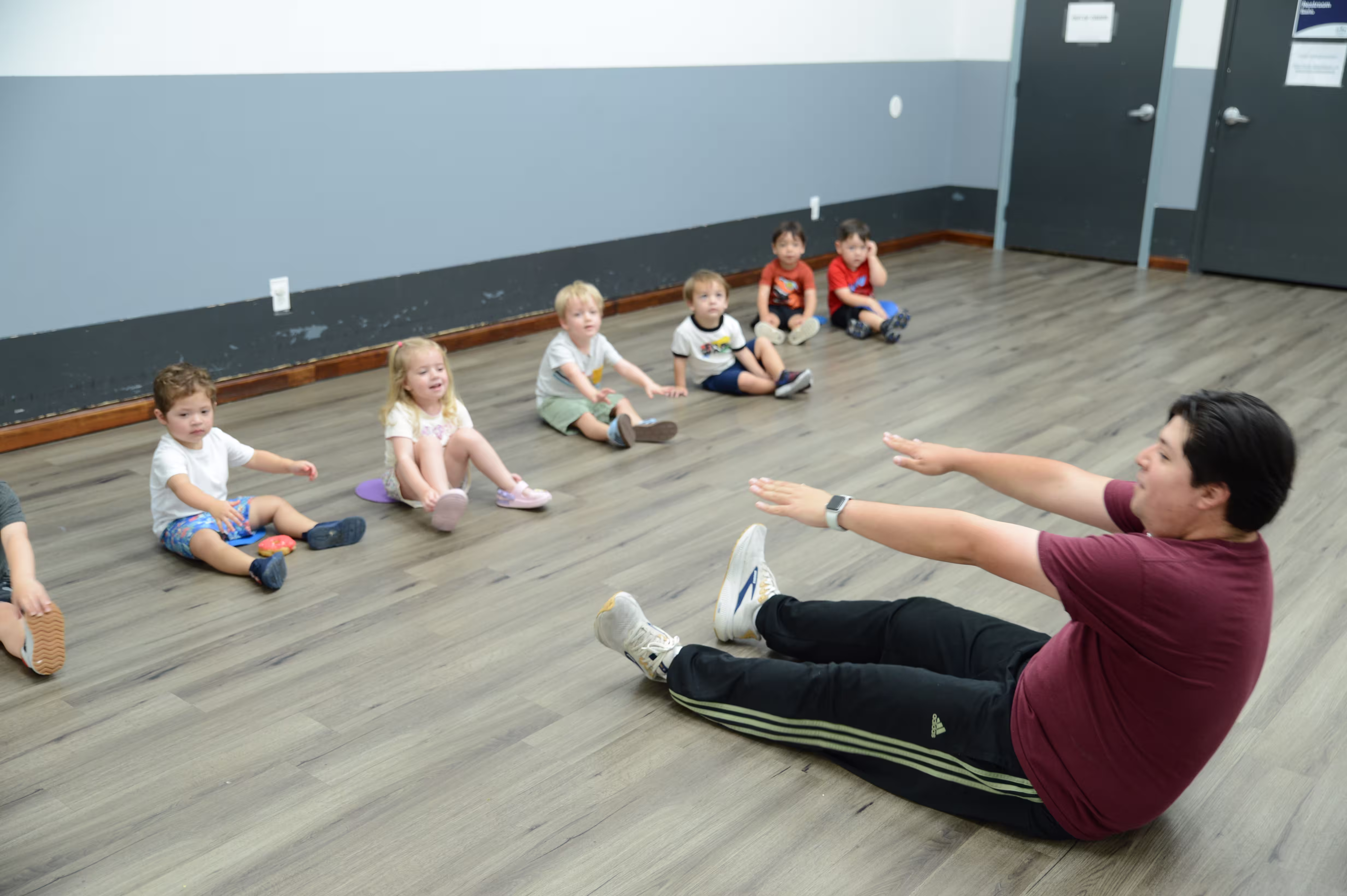 Toddlers practicing a new movement A teacher sits on the floor modeling a stretch as a group of toddlers try the same movement.