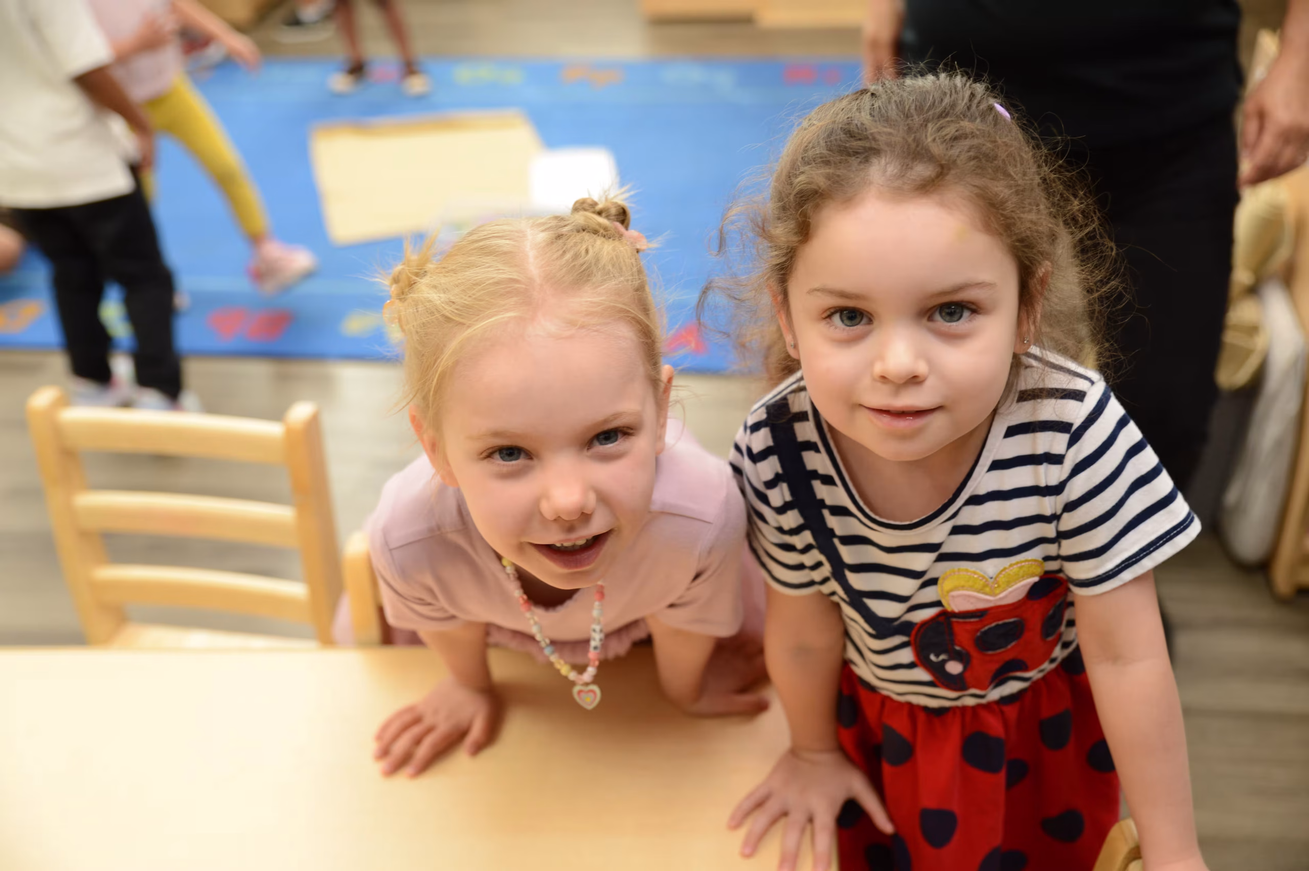 Two preschoolers in the classroom Two preschool girls lean over a classroom table and smile at the camera at Edquisitive Montessori.